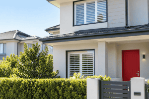 Front of a modern two-storey townhouse with a red front door, white plantation shutters and a hedge along the front fence. Article is Can a body corporate refuse renovations based on appearance?